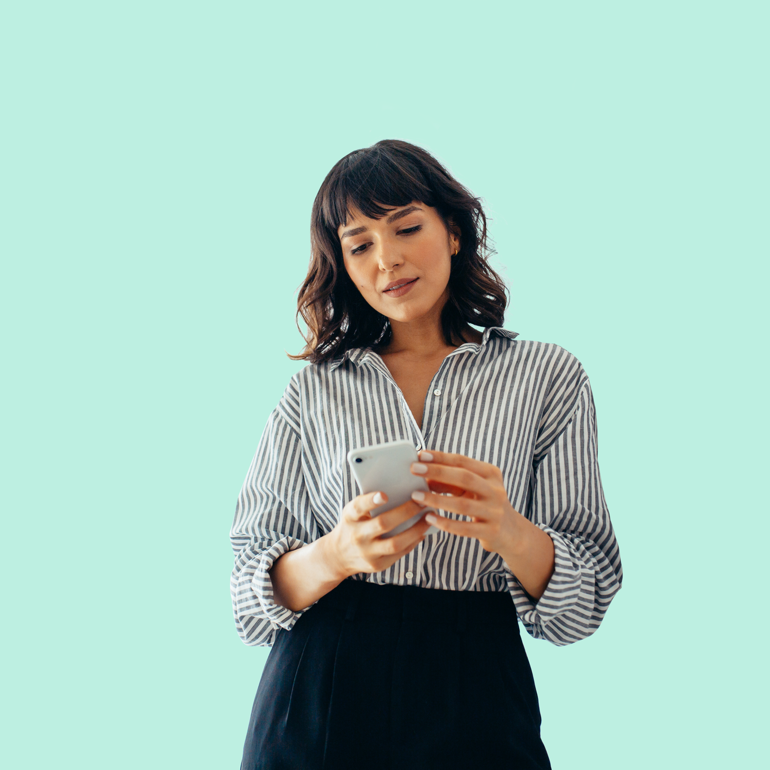 En ung kvinde med skulderlangt mørkt hår og pandehår holder en smartphone i begge hænder og kigger ned på den. Hun har en stribet skjorte og en mørk nederdel på og står op mod en ensfarvet, lyseblågrøn baggrund.